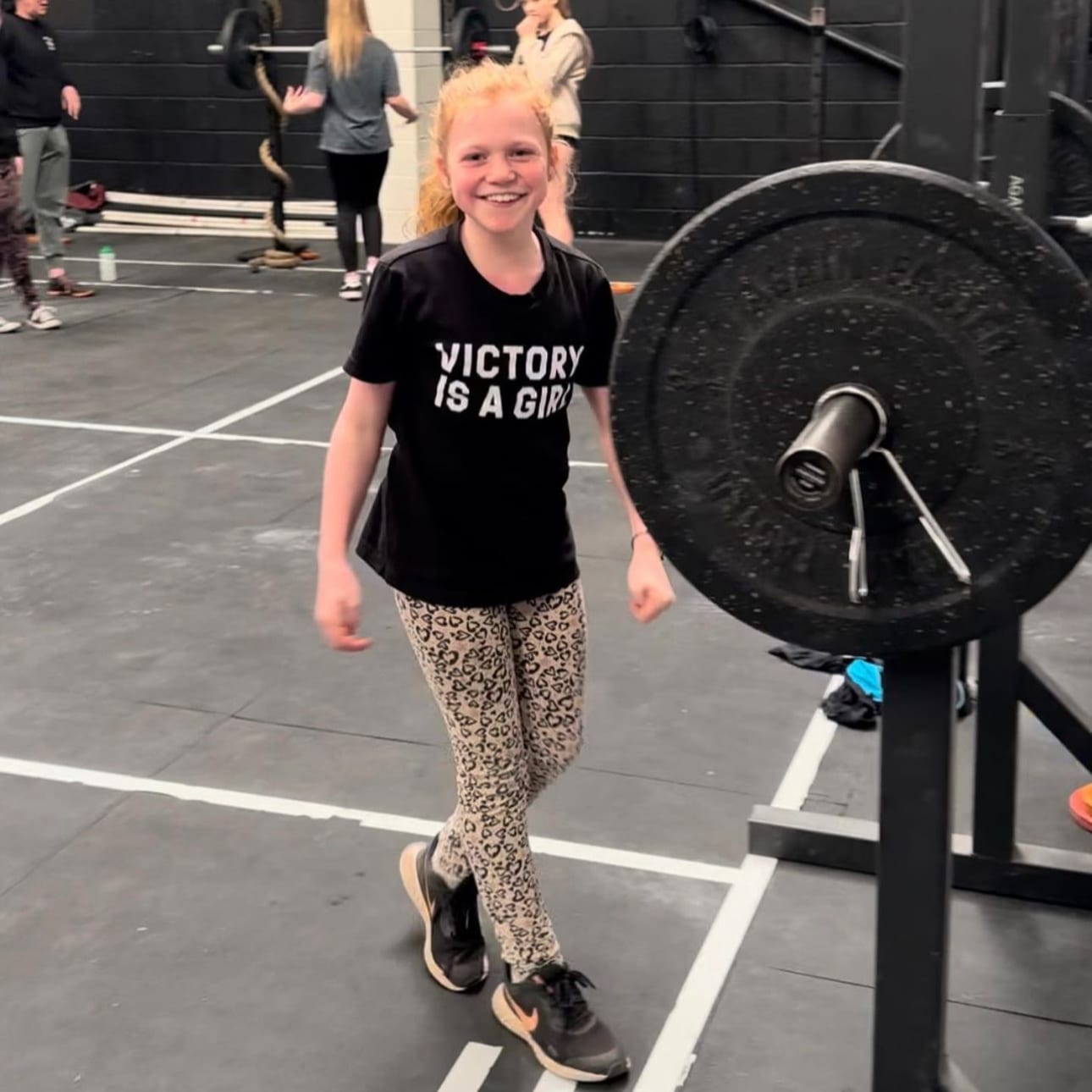 A smiling girl in a ‘Victory is a Girl’ t-shirt beside a loaded barbell